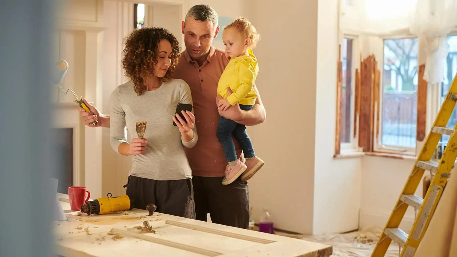 Homeowners with a young child reviewing a home renovation, with a ladder and an unfinished door on a work table